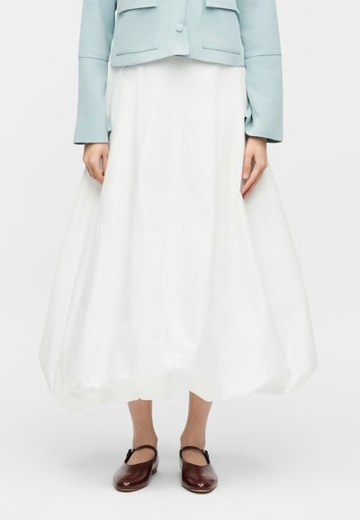 Γυναίκα που φοράει ανοιχτό γαλάζιο cropped σακάκι, λευκή φουσκωτή φούστα μήκους μέχρι το μέσο της γάμπας και σκούρα καφέ παπούτσια Mary Jane, που στέκεται μπροστά σε λευκό φόντο.