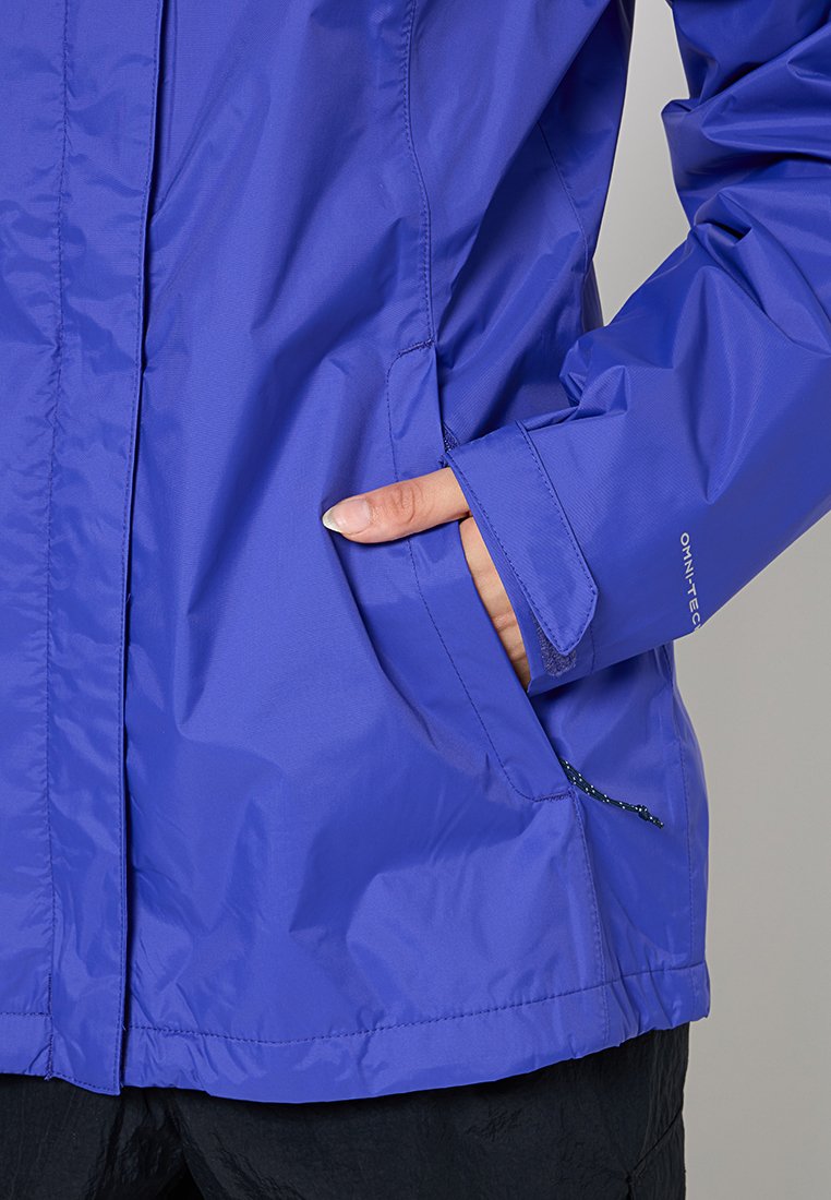 Veste violette résistante à l'eau avec une texture lisse, des poches zippées et des poignets ajustables. Design léger idéal pour le superposition.