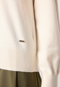 Lysbeige sweater lavet af blødt stof, med ribbede cuffs og kant. Et lille metalmærke med "Holly White" er fastgjort på siden.