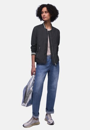 Femme aux cheveux bouclés portant une veste bomber noire, un haut rose clair, un jean bleu retroussé, des baskets argentées, tenant un grand sac cabas argenté.