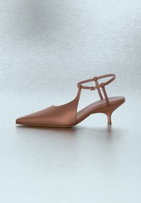 Καφέ slingback με μυτερή μύτη, τακούνι kitten, λουράκι στον αστράγαλο και μικρή αγκράφα σε λεία, αντανακλαστική επιφάνεια.