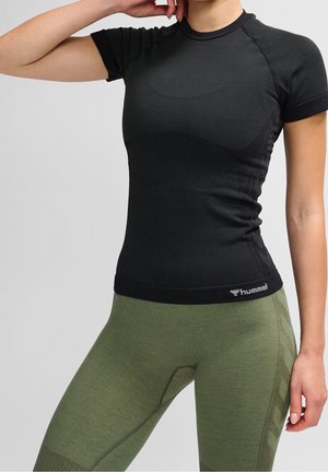 Personne en forme portant un haut de sport noir ajusté à manches courtes et des leggings texturés vert olive, posant avec un bras plié et la main près de la tête.