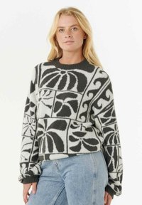 Donna bionda che indossa un maglione a fantasia bianco e nero con design floreali e geometrici astratti, abbinato a jeans azzurri chiari.