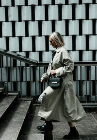 Lange, lichtgrijze trenchcoat met brede mouwen, zwarte handtas en chunky zwarte laarzen. Gelaagde muur op de achtergrond met geometrische patronen.