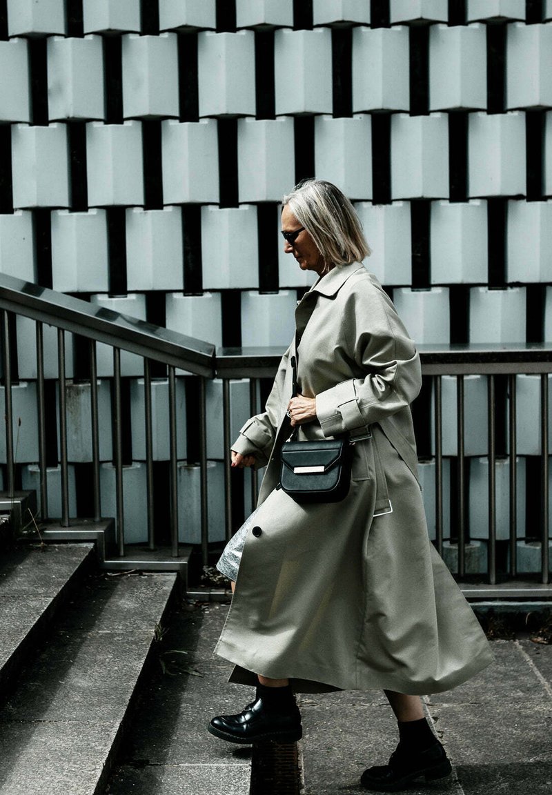 Lange, lichtgrijze trenchcoat met brede mouwen, zwarte handtas en chunky zwarte laarzen. Gelaagde muur op de achtergrond met geometrische patronen.