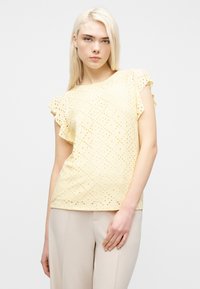 Νεαρή γυναίκα με ίσια ξανθά μαλλιά, φορούσα ανοιχτό κίτρινο μπλουζάκι με κοντά μανίκια και σκουρόχρωμο μπεζ παντελόνι, όρθια μπροστά σε ένα λευκό φόντο.