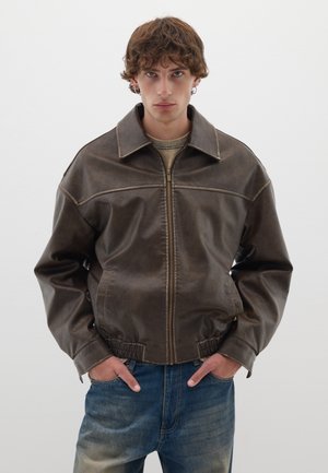 Veste en cuir brun avec un col, fermeture éclair à l'avant et taille élastique. Présente des coutures contrastantes et une coupe décontractée. Portée par-dessus un haut léger.