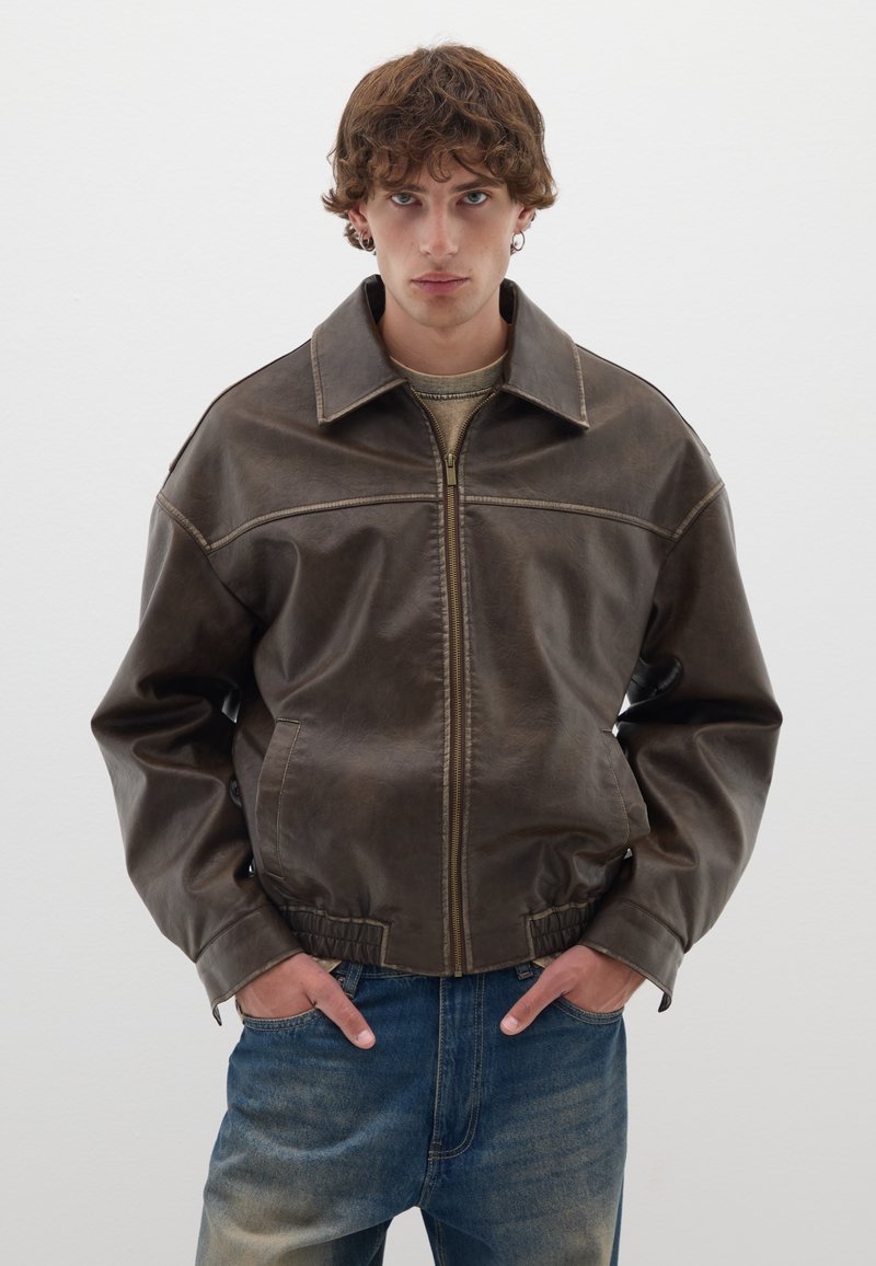 Giacca in pelle marrone con colletto, chiusura con zip e vita elastica. Presenta cuciture a contrasto e una vestibilità morbida. Indossata sopra una maglietta leggera.