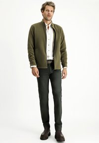Giacca in pile verde oliva con zip frontale, camicia bianca sotto, pantaloni verde scuro e scarpe marroni.