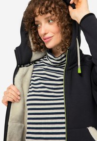 Felpa nera con zip e dettagli verdi, dotata di una morbida fodera interna. Maglione a collo alto a righe sotto, con righe grigie e blu navy.