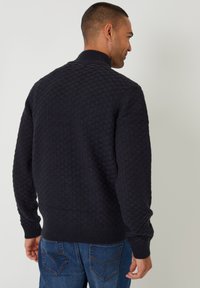 Strukturierter, dunkelblauer Pullover mit hohem Kragen und gerippten Bündchen. Das Material weist ein Karomuster auf und wird mit Jeans in Denim kombiniert.