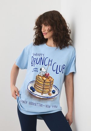 Donna con capelli ricci che indossa una maglietta azzurro chiaro con Snoopy su una pila di pancake con un testo sul brunch club di New York.