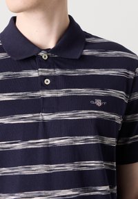 Marineblå stripet polo-skjorte med hvite, bølgende horisontale linjer, tre-knapp placket og brodert logo på brystet. Glatt bomullstekstur.