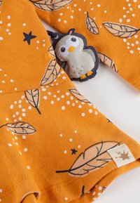 Orange fabric featuring leaf and star patterns, with a plush owl attachment and a small label. Soft texture, playful design.