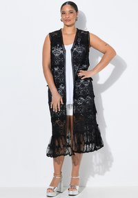 Svart spetsväst med öppen front, med intrikata blommönster och en fransad nederkant. Parat med en vit topp och silvriga plattformsskor.