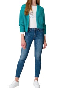 Türkisfarbener Strickcardigan mit Rippstruktur, getragen über einem weißen Grafikt-Shirt, kombiniert mit blauen Skinny Jeans und weißen Sneakers.