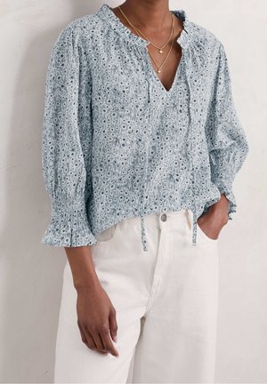 Woman wearing light blue floral blouse with smocked cuffs and white high-waist pants, standing with one hand in pocket against plain background.