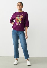 Lila übergroßes T-Shirt mit Bonbon-Graphic und Text, kombiniert mit flared blauen Jeans und weißen Sneakers auf einem neutralen Hintergrund.