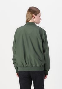 Groene bomberjack, glad fabric, elastische manchetten en zoom, ontspannen pasvorm, geen zichtbare sluitingen, gedragen met zwarte jeans, effen achtergrond.