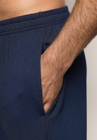 En mans hand som vilar i fickan på marinblå elastiska midjebyxor, som visar tygtextur och detaljer av armhår.