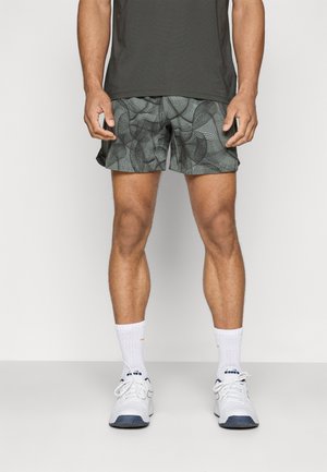 Homme portant un short de sport gris à motifs, un t-shirt noir, des chaussettes blanches montantes et des baskets blanches, debout devant un fond clair.