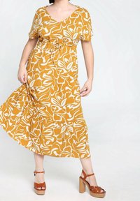 Żółta kwiatowa sukienka maxi z krótkimi rękawami, ściągnięta w talii i z falbaniastym dołem. Para brązowych sandałów na blokowym obcasie dopełnia całość.