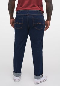 Pantaloni din denim bleumarin cu tăietură dreaptă, având două buzunare pe spate cu cusături portocalii și tivuri rulate deasupra tenișilor albi.