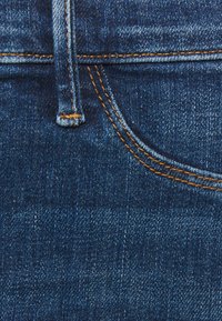 Mörkblå denimtyg med synlig vävstruktur, orange sömmar längs sömmarna och en subtil design av framfickan.