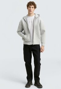 Sivá mikina na zips s kapucňou, oblečená cez biele tričko, doplnená o čierne nohavice a tmavé topánky. Model stojí na neutrálnom pozadí.