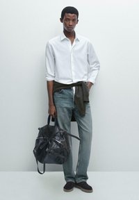 Witte bloes met knopen, donkere trui om de taille, blauwe spijkerbroek, bruine suède schoenen, met een donkere leren tas met ritsen.