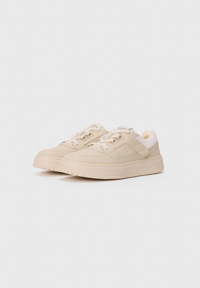 Ett par beige låga sneakers med snören och vadderade vita anklar på en vit bakgrund.