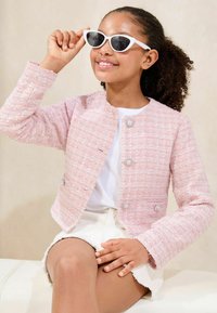 Rosa Tweed-Jacke mit rundem Kragen, zwei Knöpfen und strukturiertem Stoff. Getragen über einem weißen Oberteil und kombiniert mit stilvollen weißen Katzenaugen-Sonnenbrillen.
