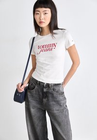 Bílý přiléhavý tričko s červeným logem "tommy jeans", doplněné volnými šedými džíny. Modelka drží malou modrou kabelku.