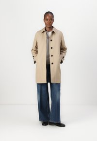 Trench-coat beige avec un col, des boutons sur le devant et deux poches latérales. Porté par-dessus une chemise grise et associé à un jean bleu à jambes larges.