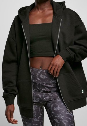 Donkere huidkleurige persoon draagt een zwart cropped tanktop, een zwarte zip-up hoodie en zwarte legging met een wit paisleypatroon, met een navelpiercing zichtbaar.