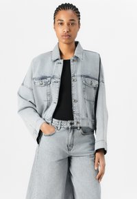 Ljusgrå denimjacka med knappstängning, två bröstfickor och en avslappnad passform. Kombinerad med matchande vida jeans och en svart topp.