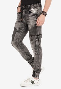 Pantalons jogger en denim gris avec des panneaux texturés, taille élastique, poignets zippés et poches avant. Portés avec des baskets blanches.