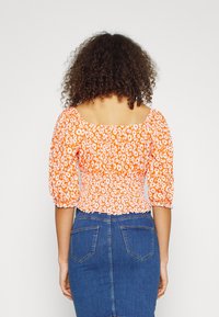Blusa floreale arancione con maniche a sbuffo, busto arricciato e scollatura quadrata, abbinata a una gonna di jeans blu con tasche posteriori.