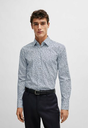 Chemise blanche avec un motif bleu marine dispersé, manches longues, col boutonné et coupe ajustée. Associée à une ceinture noire et un pantalon sombre.
