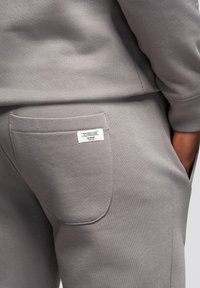 Gros plan sur la poche arrière d'un jogging gris avec une étiquette cousue sur laquelle est inscrit "Your only limit is your mind" et une main dans la poche latérale.