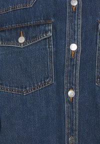 Jeansskjorta med mörkblå tvätt, silverfärgade tryckknappar, två bröstfickor och kontrasterande gula sömnadsdetaljer.