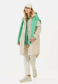 Beige Regenmantel mit grüner Kapuze, über einem grünen Shirt und cremefarbenen Hosen getragen. Helle Sneakers runden das Outfit ab.