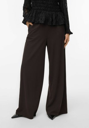 Donna che indossa pantaloni larghi scuri, scarpe con punta nera e una maglietta nera a maniche lunghe con texture, con una mano in tasca.