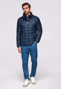 Marineblauer Steppjacke mit horizontalen Nähten, hohem Kragen und Reißverschluss; getragen über blauen Jeans und weißen Sneakers.