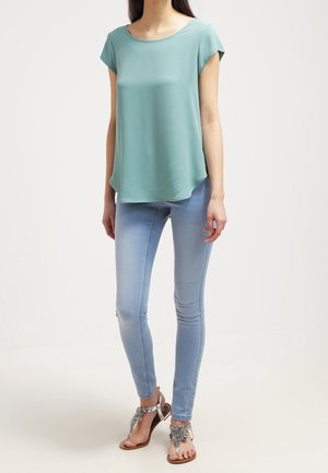 Femme portant un haut vert clair à manches courtes, un jean skinny bleu clair et des sandales argentées embellies, debout devant un fond uni.