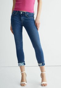 Mujer vestida con jeans ajustados oscuros arremangados con puños de estampado floral, top sin mangas rosa y sandalias de tacón blanco, de pie contra una pared blanca.