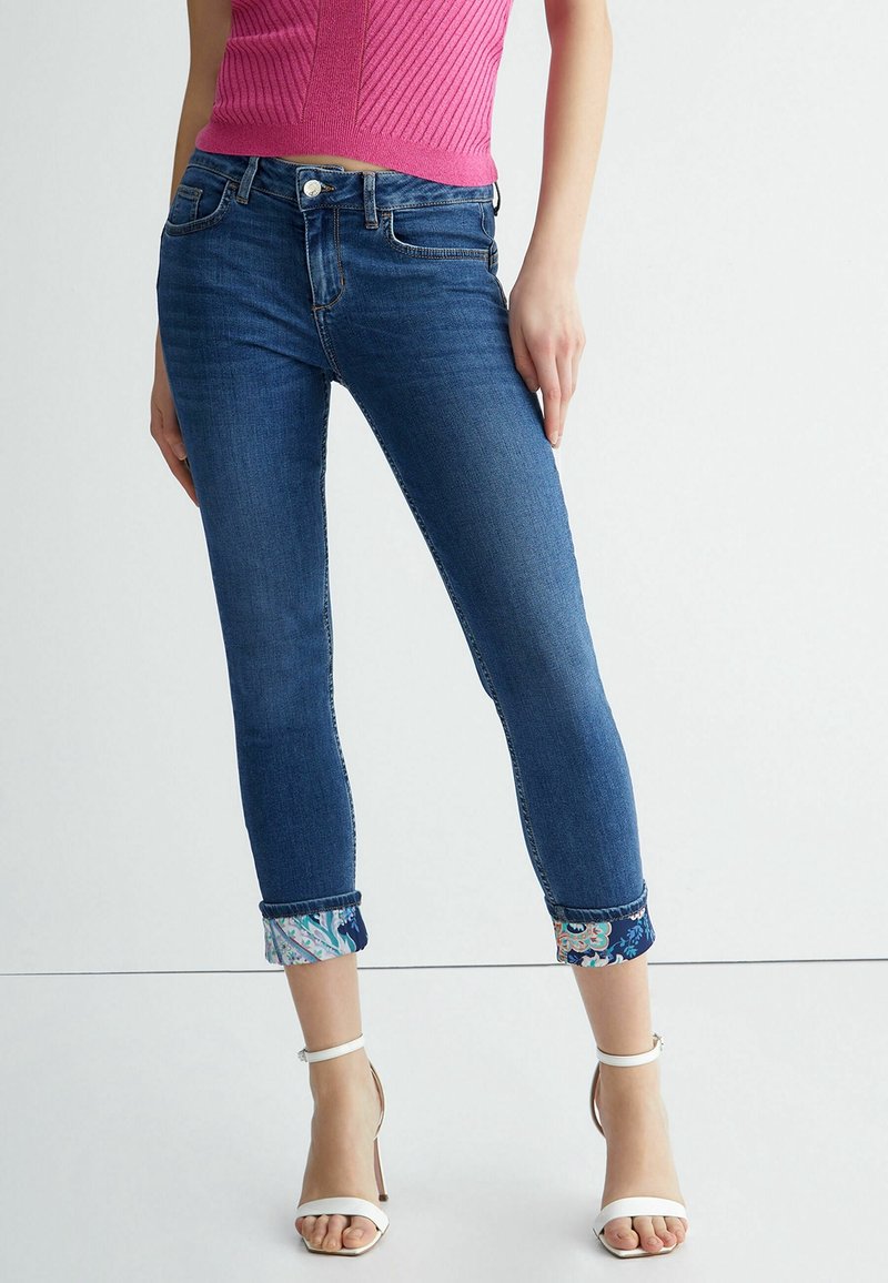 Mujer vestida con jeans ajustados oscuros arremangados con puños de estampado floral, top sin mangas rosa y sandalias de tacón blanco, de pie contra una pared blanca.