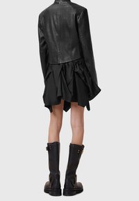 Personne portant une veste en cuir noire, une jupe noire à volants et de hautes bottes noires, vue de dos sur un fond gris.
