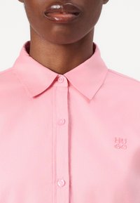 Camicia button-up rosa chiaro realizzata in tessuto morbido. Presenta un colletto classico, due bottoni e il logo ricamato "HUGO" sul lato sinistro del petto.