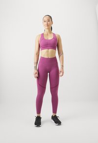 Sujetador deportivo ajustado en púrpura y leggings con tejido texturizado. Los leggings presentan detalles en paneles laterales. Zapatillas deportivas negras sobre un fondo blanco.